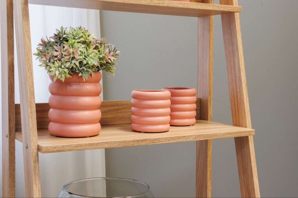 A imagem ilustra uma estante escada, na cor amadeirado claro, com vasos de cerâmica na decoração.