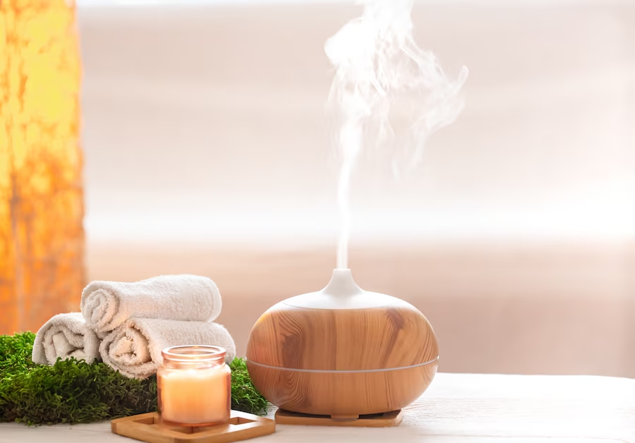 A imagem ilustra o que é aromaterapia através de um difusor de ambientes ligado. Ao lado dele encontra-se uma vela e três toalhas brancas enroladas.