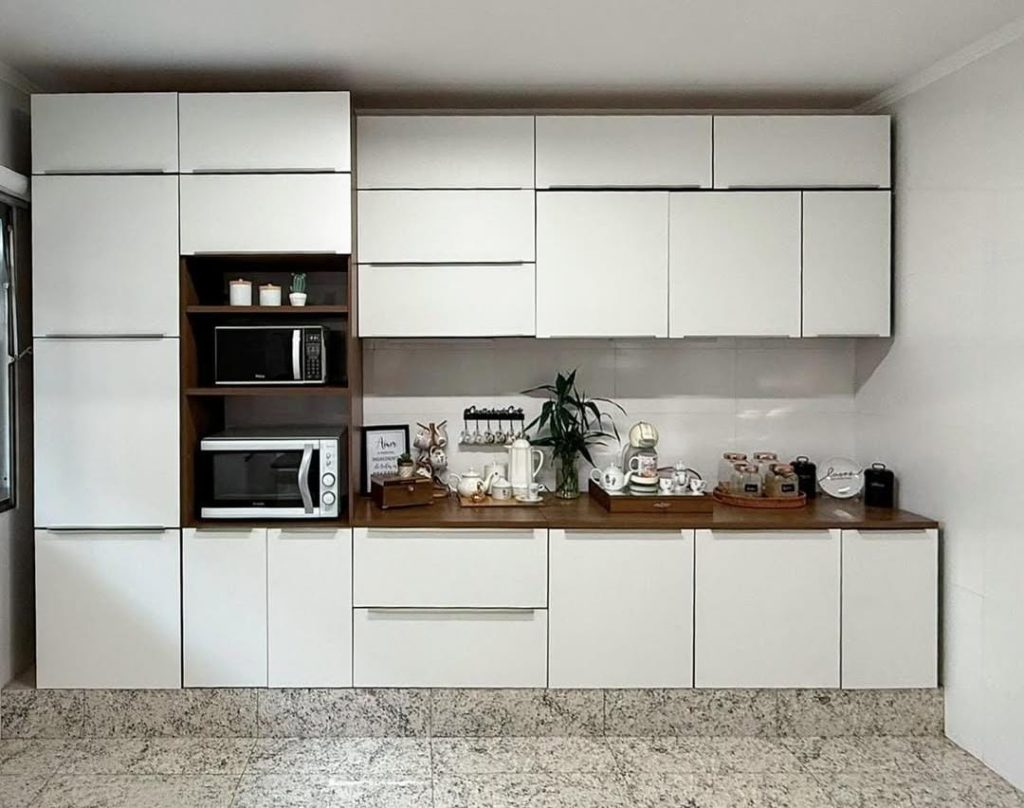 Cozinha com armários brancos, incluindo uma área central com nicho de madeira que contém micro-ondas e outros eletrodomésticos, e uma bancada de madeira com utensílios, plantas e itens decorativos.