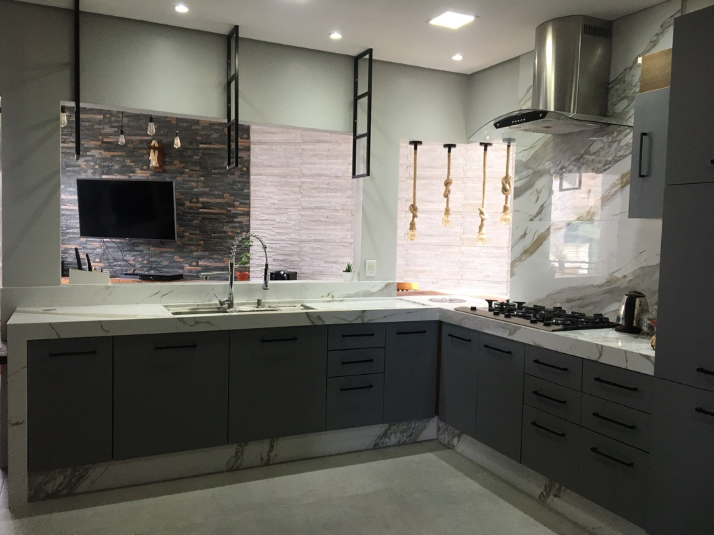 A imagem ilustra uma cozinha americana planejada na cor cinza escuro com uma pedra de mármore branco em cima e detalhes metalizados e pretos, como a pia e a geladeira.