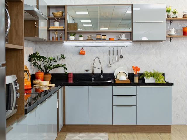 Cozinha com armários cinza-claro e detalhes em madeira, bancada preta, pia de aço inox, prateleiras abertas com utensílios e plantas, decorada com diversos objetos laranja.