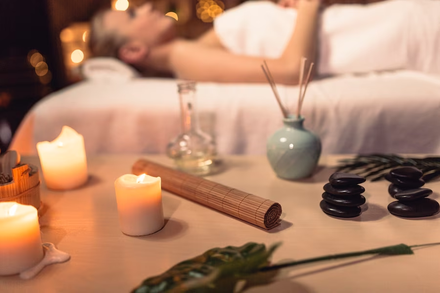 A imagem ilustra o que é aromaterapia com uma mulher deitada em uma maca de massagem ao fundo e velas sobre uma mesa à frente com essencias.