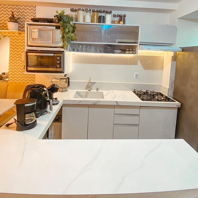 A imagem ilustra uma cozinha americana planejada, com balcões e armários brancos e eletrodomésticos à mostra, como cooktop, cafeteira e air fryer.