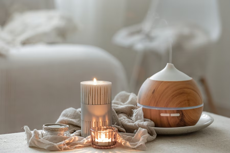 A imagem ilustra o que é aromaterapia através de um quarto branco com um difusor de essencias e velas sobre uma mesa à frente da foto. 