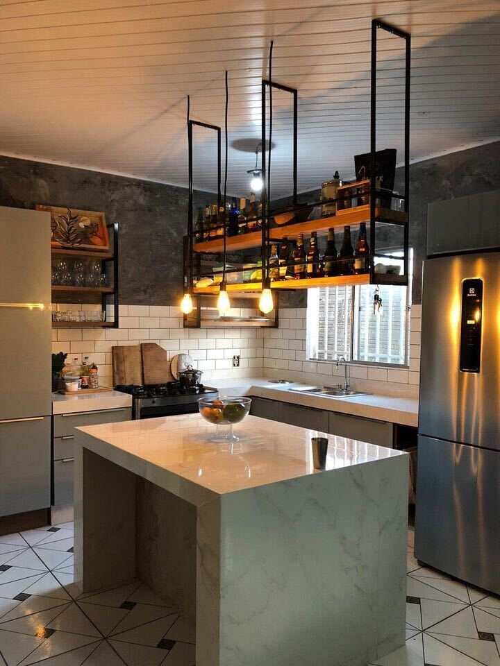 A imagem ilustra uma cozinha planejada americana com um balcão de mármore no centro. Acima dele há uma prateleira suspensa com bebidas e luminárias.