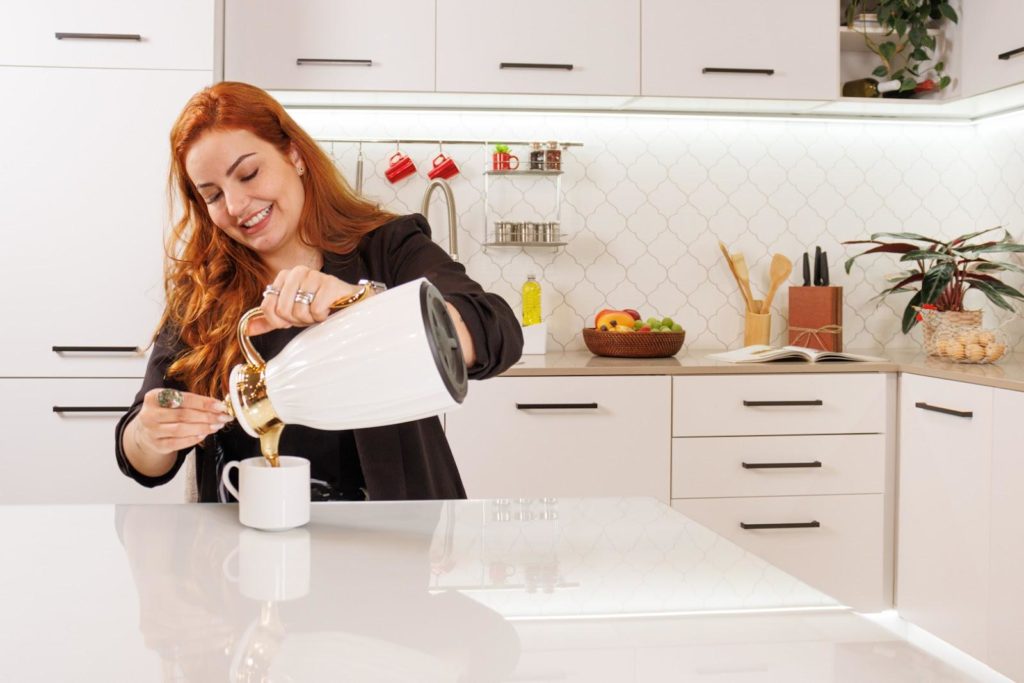 Mulher ruiva, sorrindo, colocando café numa xícara disposta no balcão de uma cozinha Agata, na cor branca.