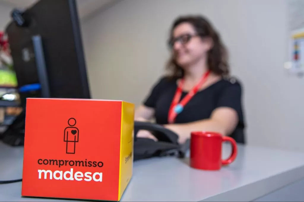 Caixa vermelha com os dizeres “compromisso Madesa” em destaque. Ao fundo, uma funcionária Madesa trabalhando, na frente de um computador.