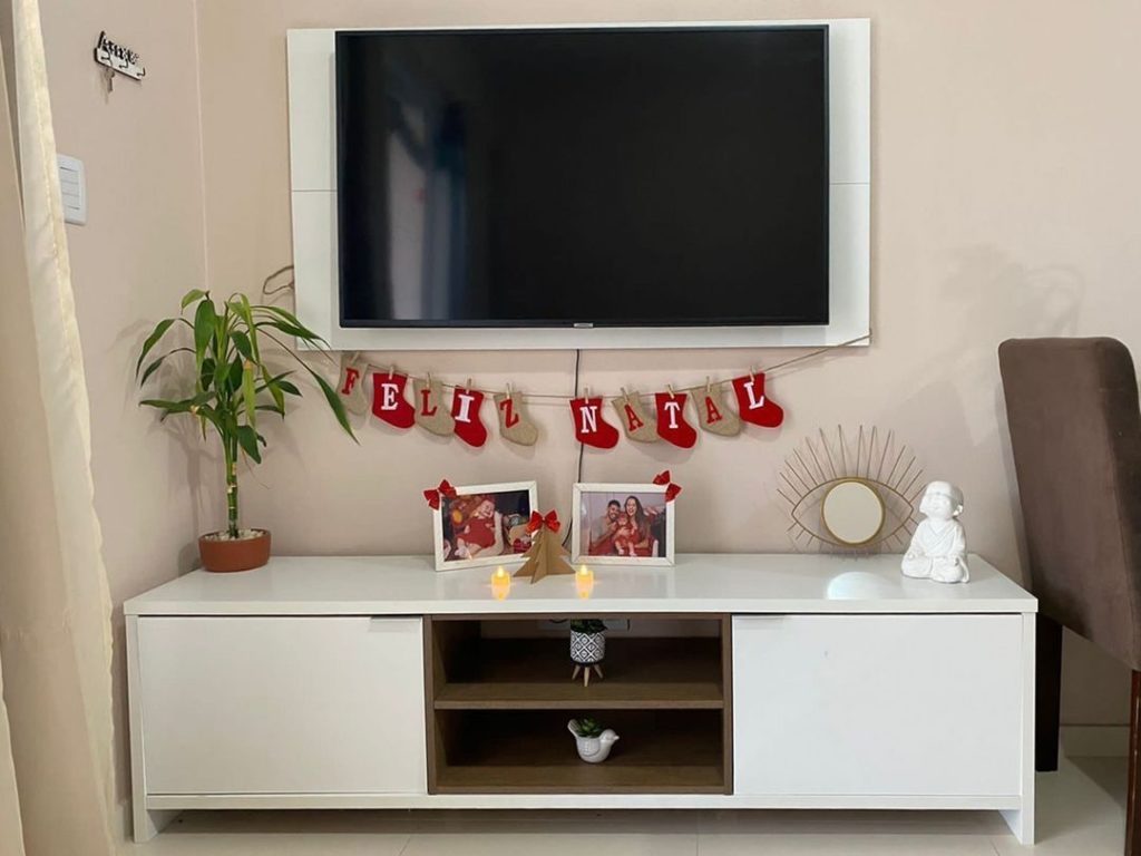 Sala de TV branca, com rack Madesa na mesma cor, com fotos de família, velas e meias de Natal penduradas escrito “Feliz Natal”