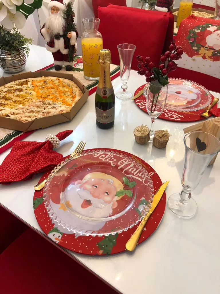 Mesa posta de Natal, com sousplat vermelho estampado, pratos transparentes, taças, vinho e outras decorações.