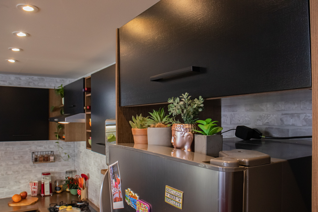 Cozinha Agata, nas cores rustic e preto, decorada com utensílios e eletrodomésticos. Em cima da geladeira, há alguns vasos de plantas decorativos.