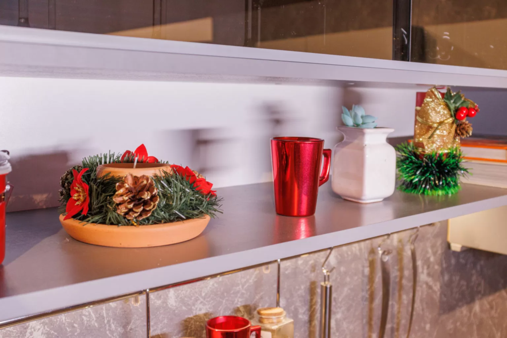 Prateleira de cozinha decorada com copos vermelhos, vasos com plantas, pinhas e outros itens natalinos.