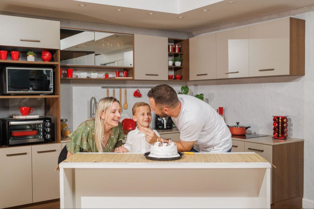 Família feliz, com a mãe, pai e filho sorrindo em frente a um bolo de aniversário branco. Ao fundo, uma cozinha Madesa na cor crema, decorada com utensílios vermelhos.