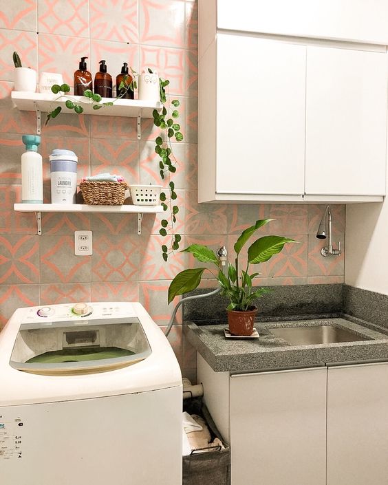 uma lavanderia está em foco, com uma máquina de lavar e armários Madesa brancos compondo seu espaço. Em cima da pia está um vaso com plantas e, na parede, estão aplicados azulejos cinza com detalhes em rosa salmão pastel. 