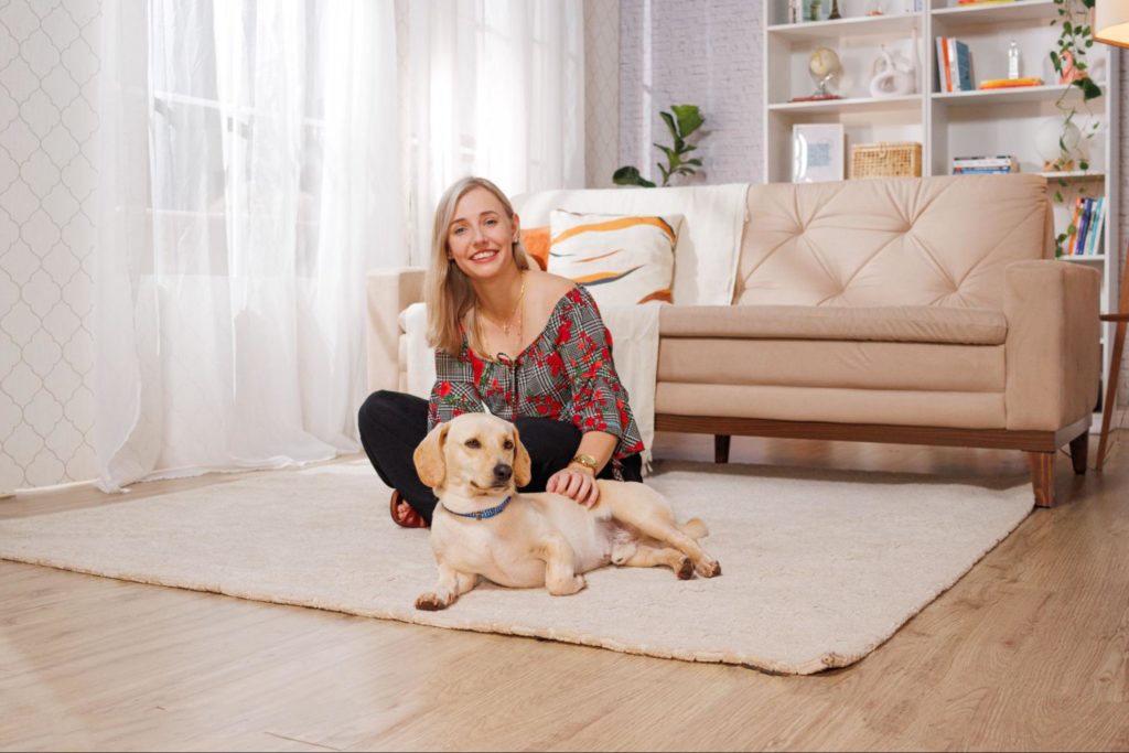 Mulher loira, sorrindo, sentada ao lado de um cachorro numa sala de estar de cores neutras e tonalidades branco e bege.