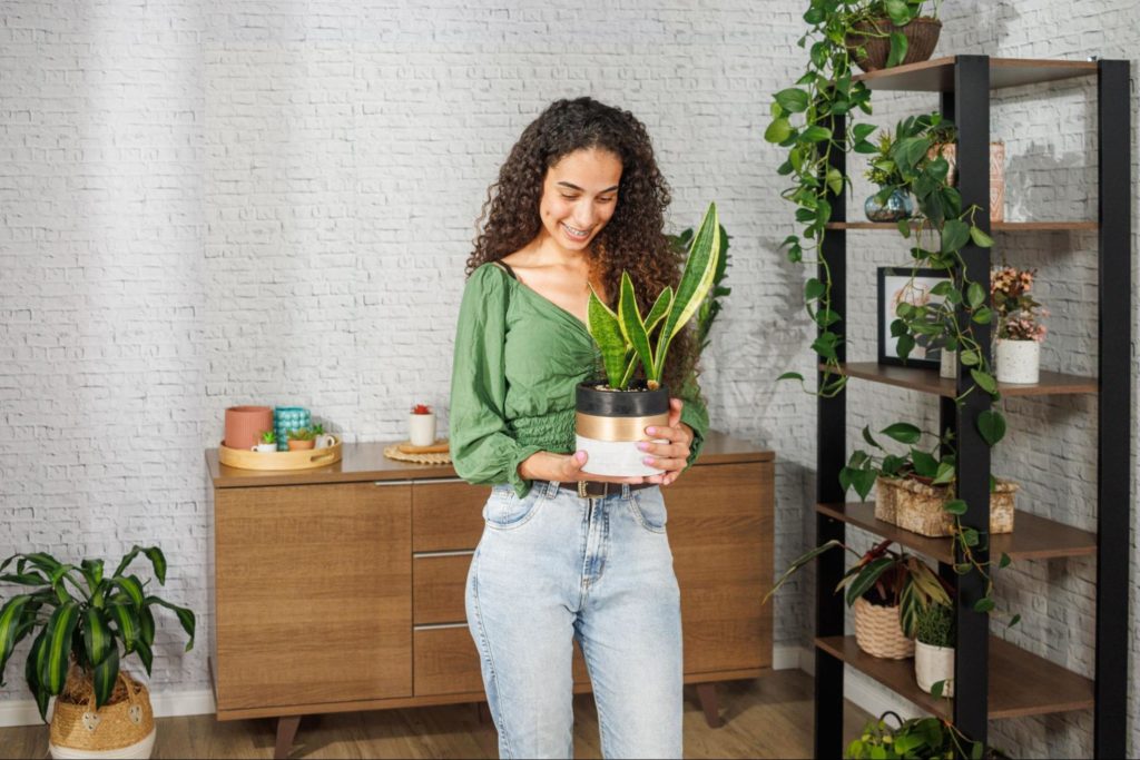 Mulher segurando uma planta nas mãos, sorrindo. Ao fundo, uma estante Madesa decorada com diversas plantas, e ao fundo um aparador Madesa.