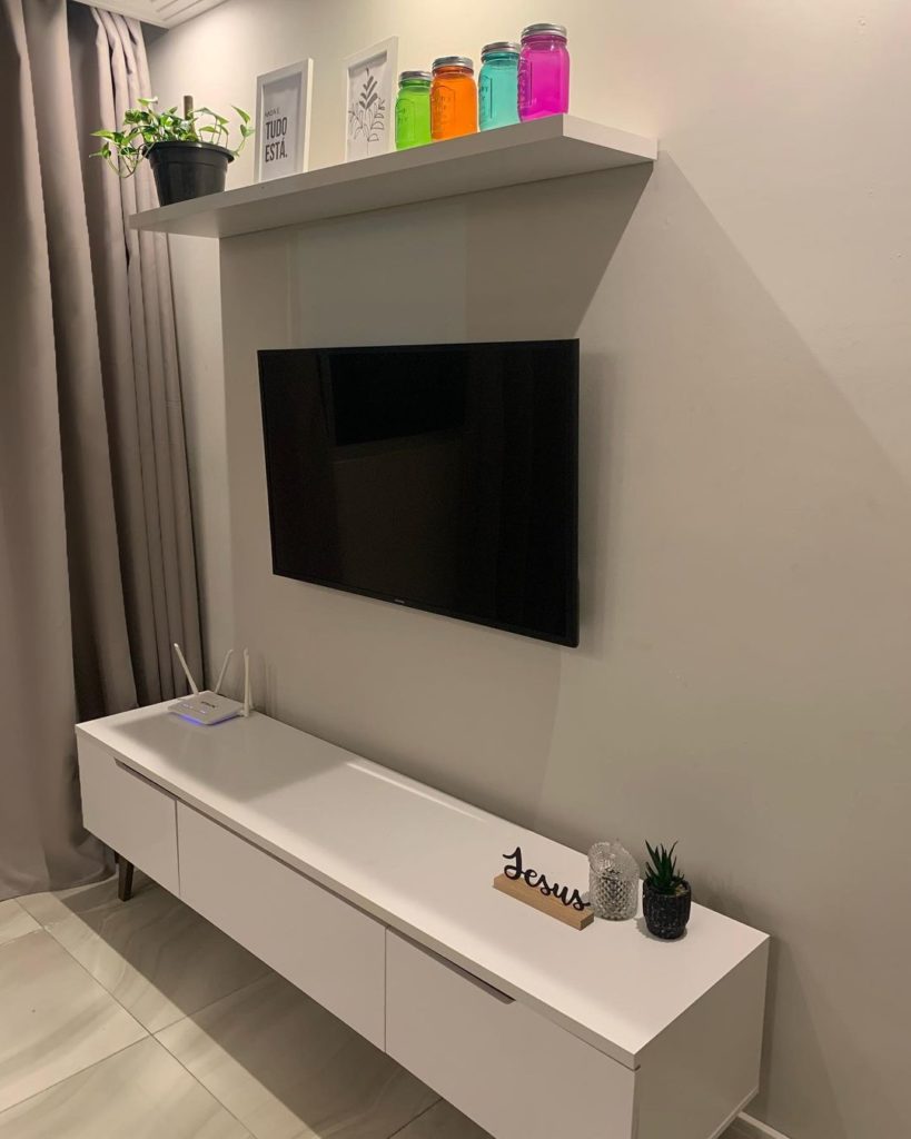 Sala de estar com rack Reims Madesa, na cor branca, com decoração minimalista com TV e uma prateleira próxima ao teto.