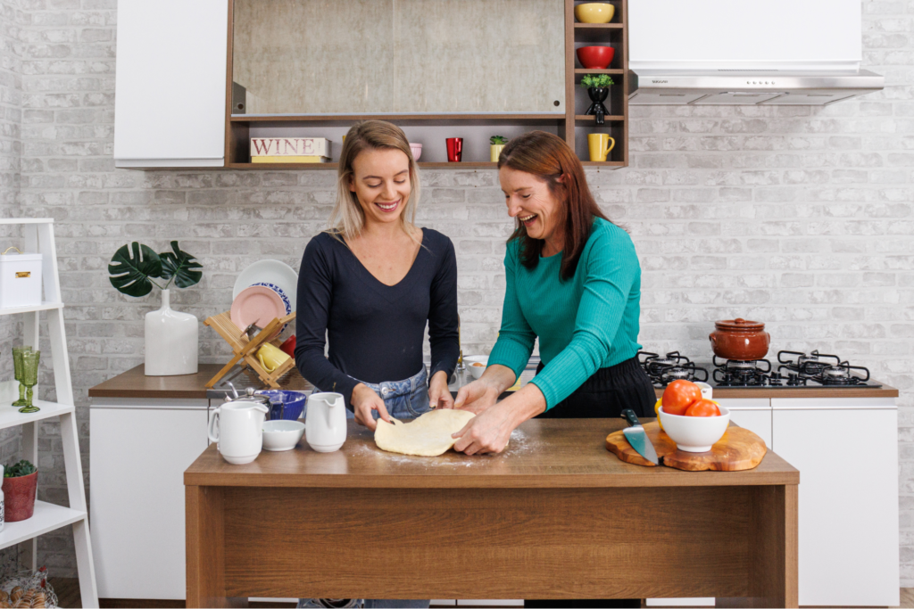 Duas mulheres estão na cozinha, sorrindo enquanto trabalham juntas na massa. A mulher à direita, com cabelo longo e castanho, veste uma blusa verde e está segurando a massa com as mãos. A mulher à esquerda, com cabelo curto e loiro, veste uma blusa preta e observa a outra. O ambiente é moderno, com prateleiras e utensílios de cozinha visíveis ao fundo, além de alguns tomates e uma panela no balcão.