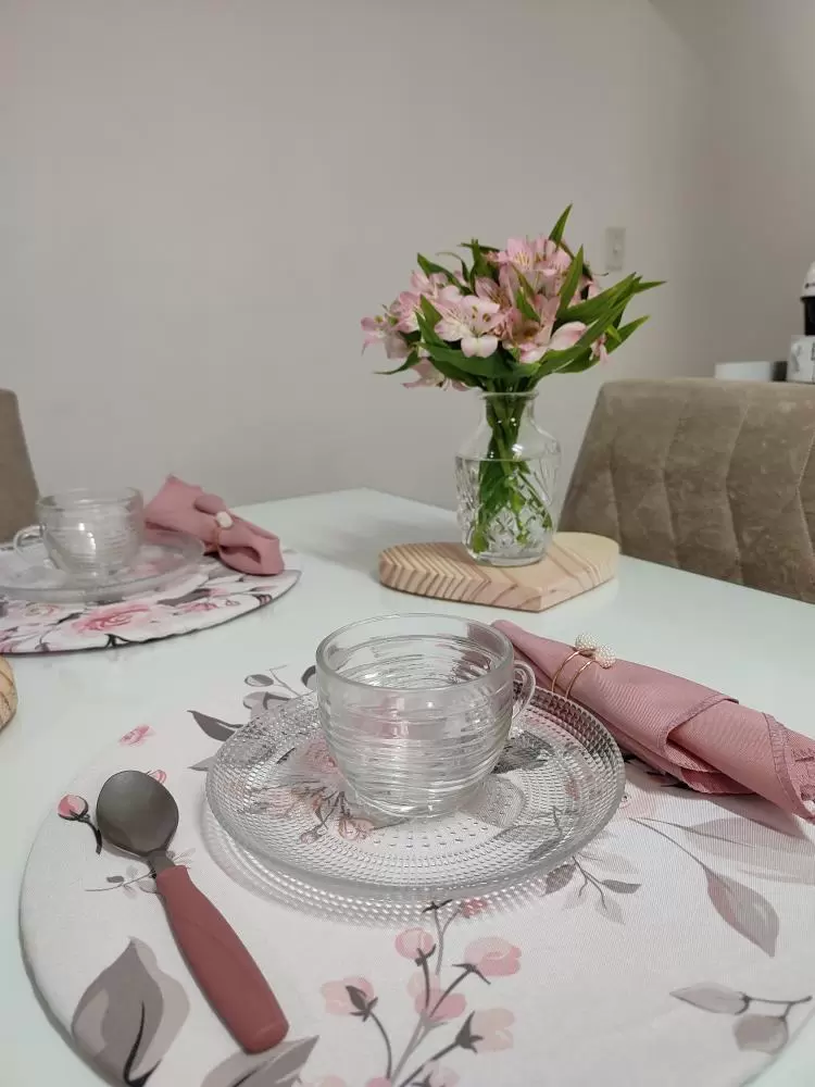 Uma mesa elegante está decorada com detalhes delicados. Um arranjo de flores rosas está em um jarro de vidro, ao fundo. À frente, um lugar está montado com um prato de vidro, uma xícara transparente e um guardanapo rosa dobrado. Um colher com cabo rosa está à esquerda do prato, e a toalha de mesa apresenta um padrão floral em tons suaves. O cenário transmite um ar acolhedor e bem cuidado.