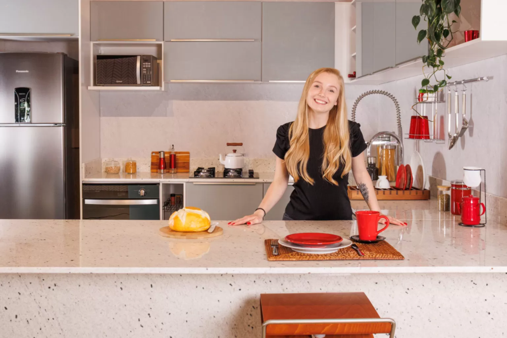 Jovem loira sorrindo, em frente a uma ilha de cozinha Madesa. Ao fundo, módulos, pia e eletrodomésticos da cozinha.