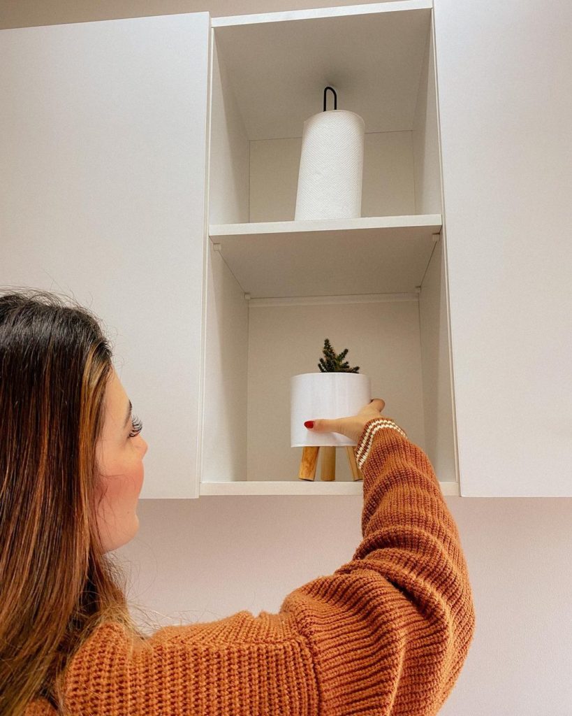 Mulher segurando um vaso de plantas decorativo, ajeitando-o em um nicho de armário de cozinha.