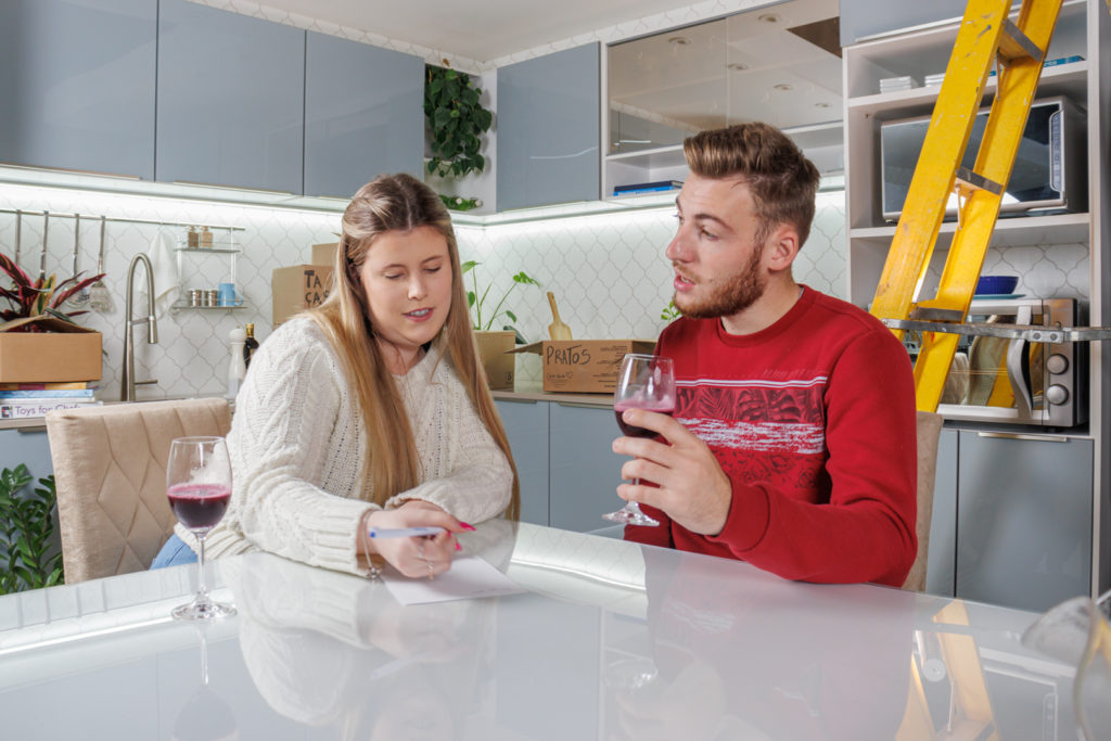 Um casal está sentado à mesa Madesa que está presente em uma cozinha grande planejada. Eles tomam taças de vinho e estão fazendo anotações enquanto conversam sobre a rotina da casa.