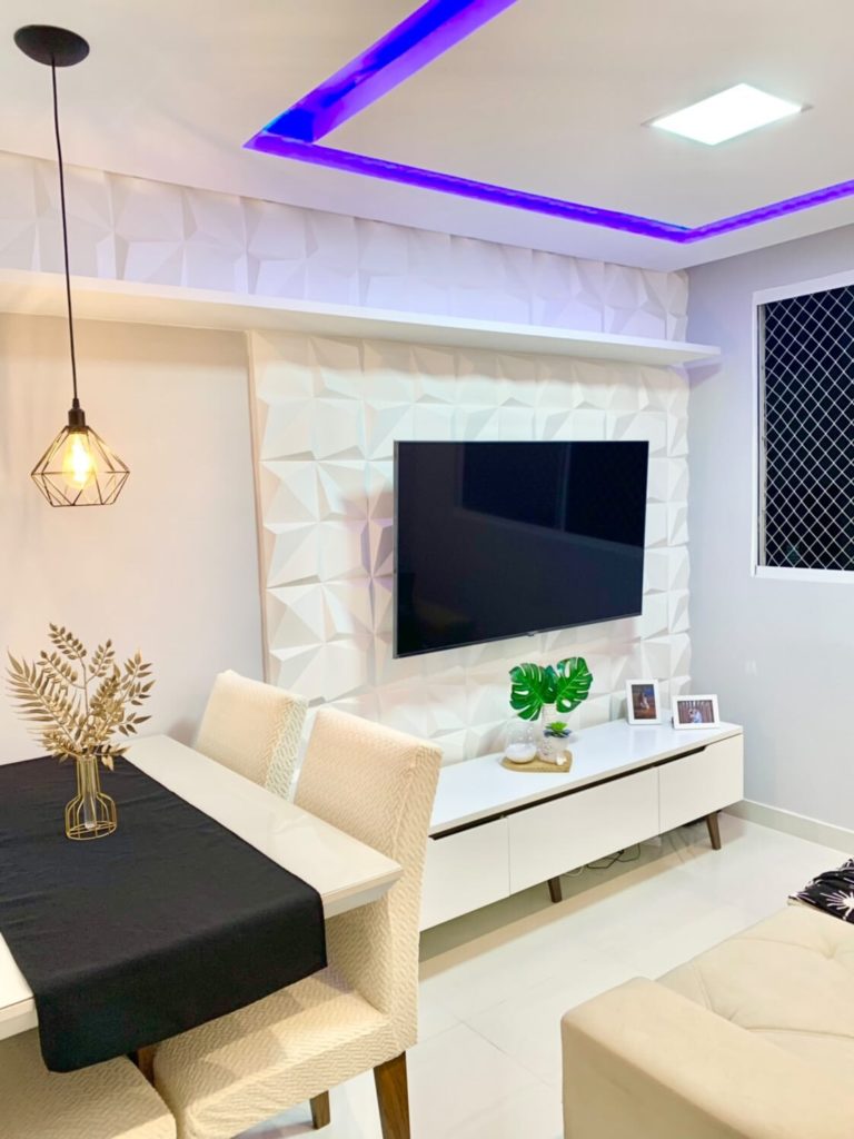 uma sala de estar com paredes claras e com detalhes em gesso tem sua iluminação colorida através de fitas de LED azul. Nela está um rack Madesa Reims branco, uma mesa com cadeiras em tons creme, uma luminária sobre a mesa e objetos de decoração dispostos pelo ambiente.