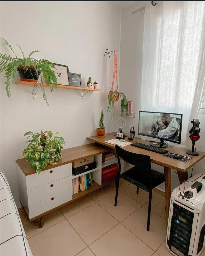  Escrivaninha Madesa Rustic Lisboa está disposta no canto de um quarto. Em sua superfície estão dispostos um monitor, vasos com plantas e objetos de escritório. 