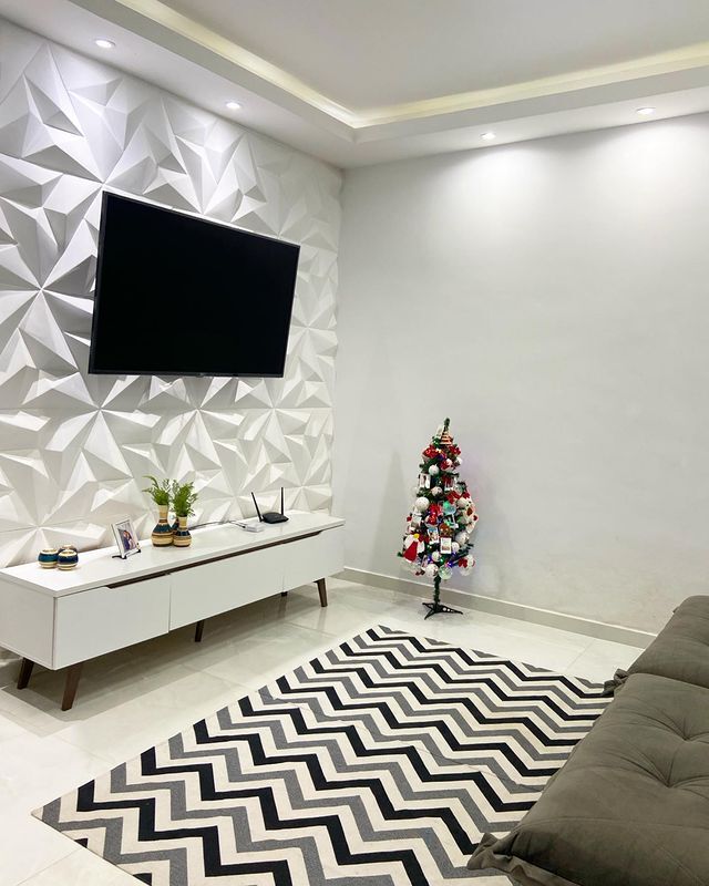 uma sala com paredes brancas e detalhes em gesso tem sua iluminação vinda do teto, dividida por spots de luz. Seu tapete é branco e preto em formas geométricas e, no canto do ambiente, está uma árvore de natal pequena.