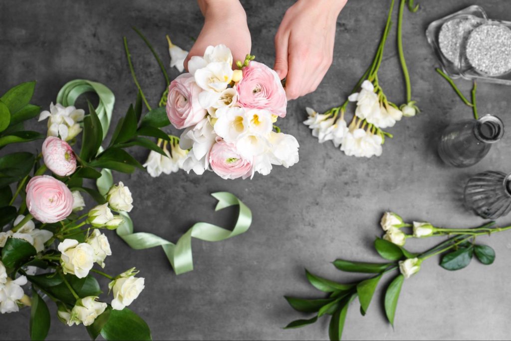 Duas mãos femininas estão segurando ramos de flores creme e cor de rosa. Ao fundo, está uma superfície cinza que tem outros ramos de folhas e flores espalhados, além de pequenos vasos de vidro e uma fita.