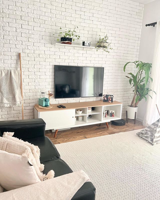 uma sala com parede de tijolos brancos está decorada com um tapete creme, um rack Madesa Dubai, um vaso de planta e uma TV.