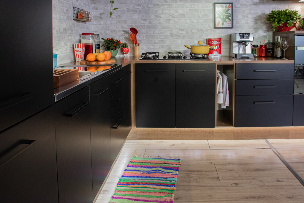 Uma cozinha Madesa Ágata preta está em um ambiente amplo, e acompanha o movimento da parede em L. Há um tapete colorido no chão e, sobre os balcões da cozinha, estão dispostos diversos eletrodomésticos, alimentos e utensílios de cozinha.
