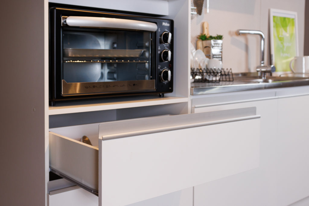 Um forno está acoplado na cozinha Madesa Topázio Branca enquanto seu gaveteiro está aberto logo abaixo. Ao fundo é possível notar um balcão da cozinha que contém utensílios em sua superfície.