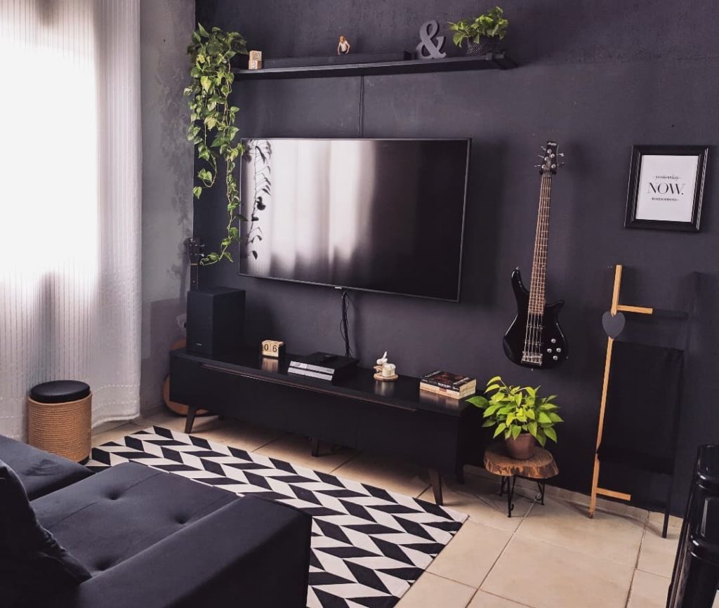 uma sala com paredes pretas está decorada com sofá escuro, tapete branco e preto com formas geométricas, vasos de plantas e uma guitarra, acoplada do lado esquerdo da televisão.