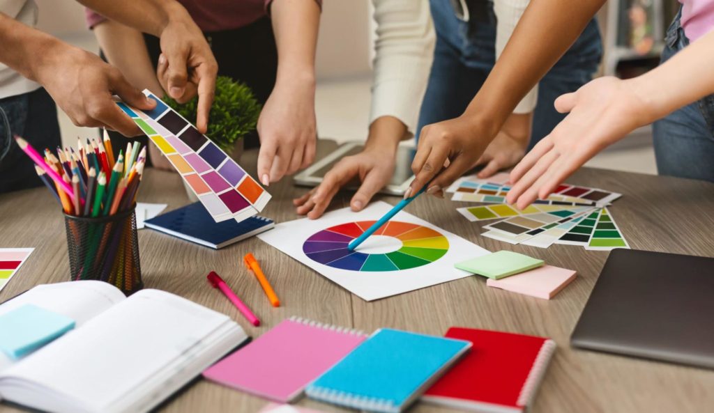 as mãos de diversas pessoas estão sobre uma mesa com cartilhas de cores e um círculo cromático impresso. Alguns deles seguram algumas cartelas de cores na mão enquanto outros seguram canetas e lápis, apontando para o círculo cromático na hora de fazer combinações.