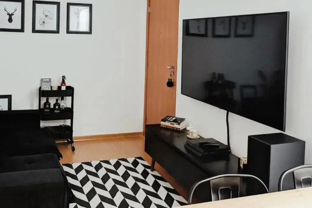 Sala de estar com sofá preto. Na parede, há quadros emoldurados com ilustrações. Uma televisão grande está montada na parede, acima de um móvel escuro com objetos decorativos. 