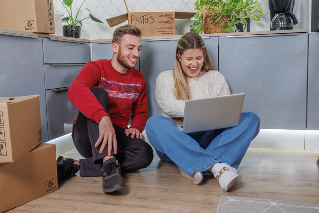 Casal sentado no chão de uma cozinha enquanto olham oportunidades de móveis na black friday no notebook. A cozinha conta com armários Madesa cinza e caixas de mudança