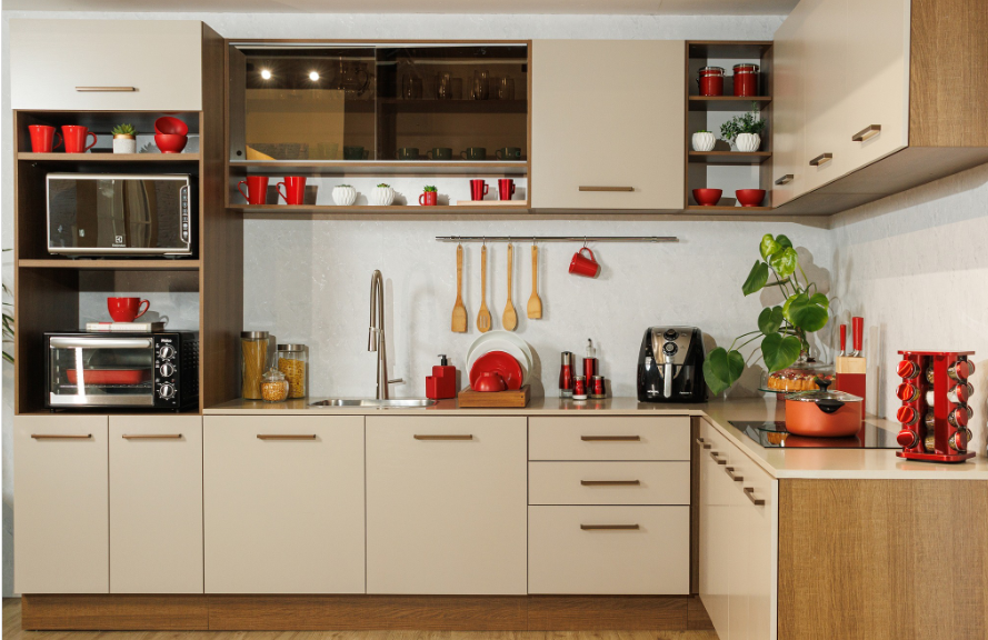 cozinha em L com armários de portas claras e detalhes em madeira. Utensílios com detalhes em vermelho dispostos sobre sua superfície e prateleiras.