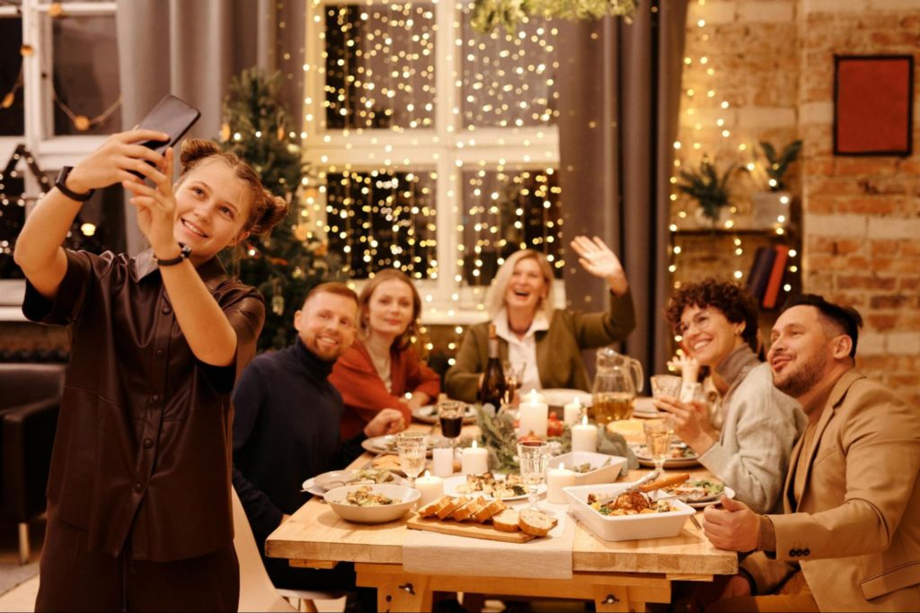 família reunida em mesa de ceia de natal. Menina segura celular e tira selfie com as pessoas sentadas na mesa, que sorriem e olham para a foto.