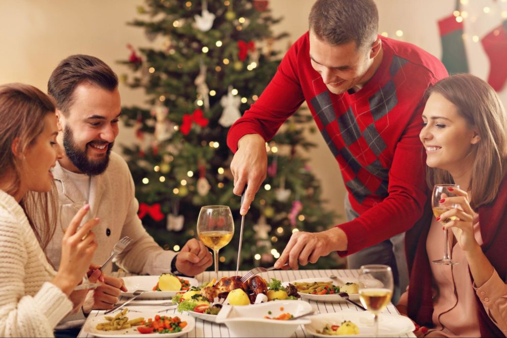 pessoas reunidas ao redor de ceia de natal, com árvore natalina e decorações temáticas ao fundo