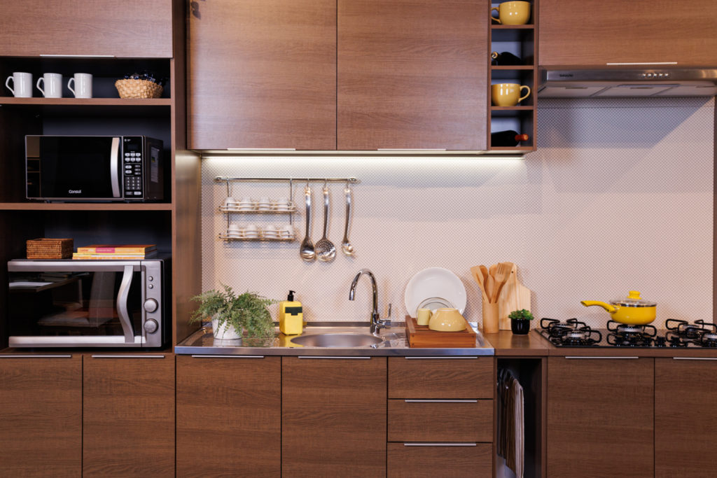 cozinha Madesa com armários em madeira tem torre quente com microondas e forno, e utensílios com detalhes em amarelo dispostos pelo balcão, além de um vaso de plantas, xícaras e um cooktop