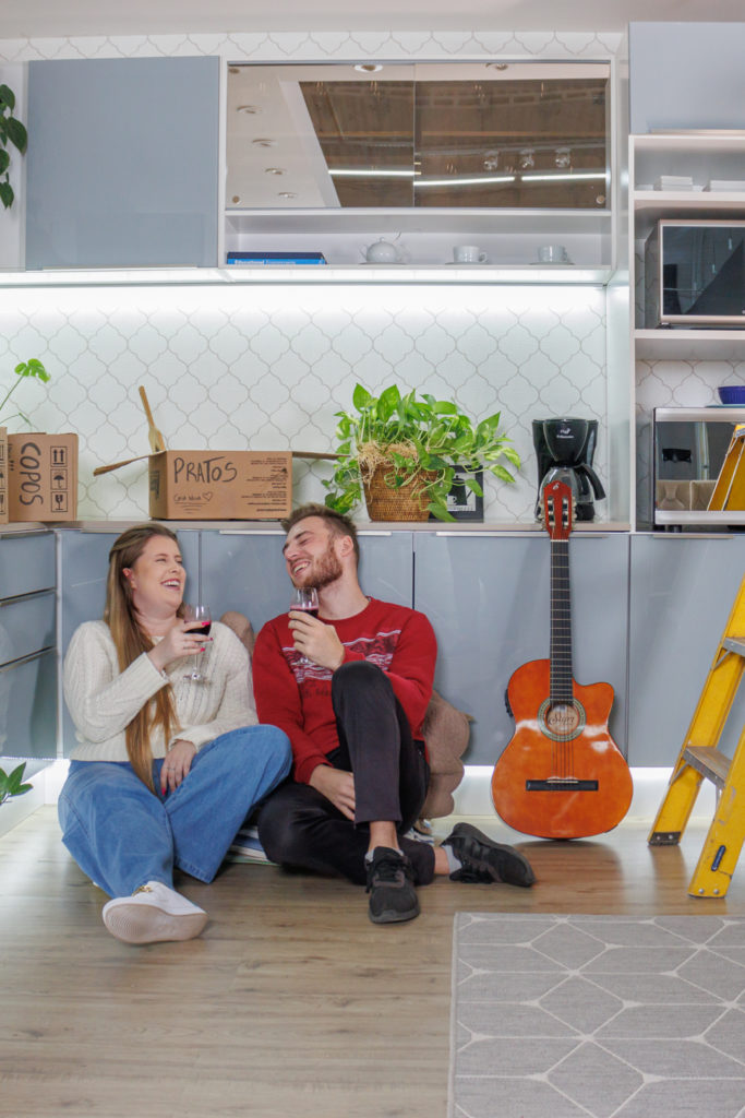 casal sentado no chão de cozinha com armários madesa cinza e chão claro. Sorriem e seguram taças de vinho ao lado de uma escada de serviço, um violão e caixas de papelão típicas de mudança, com o nome de utensílios domésticos.