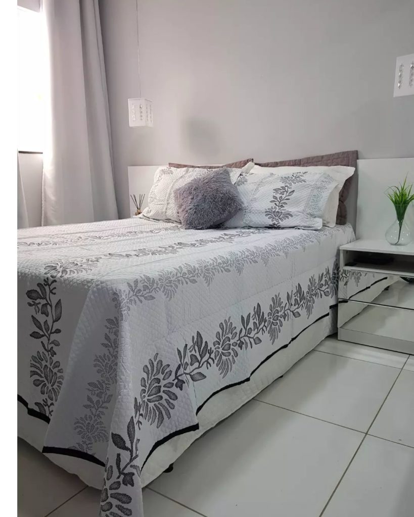 quarto com piso frio e janelas abertas tem cama de casal arrumada com colchas claras e almofadas na mesma tonalidade