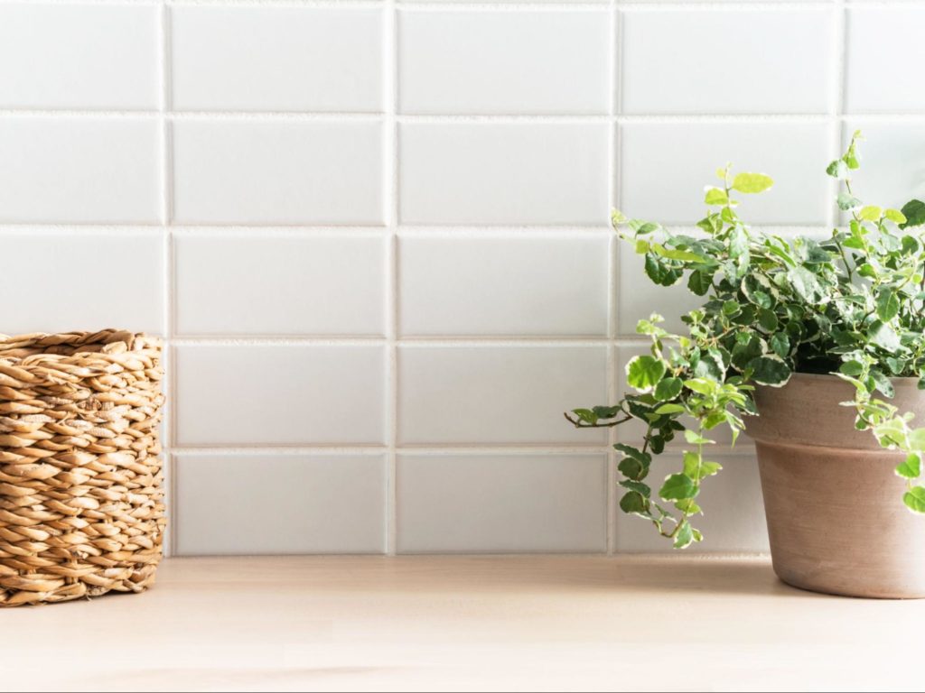  vaso de plantas ao lado de cesta  de vime, dispostas sobre mesa de madeira. Azulejos brancos na parede de fundo.