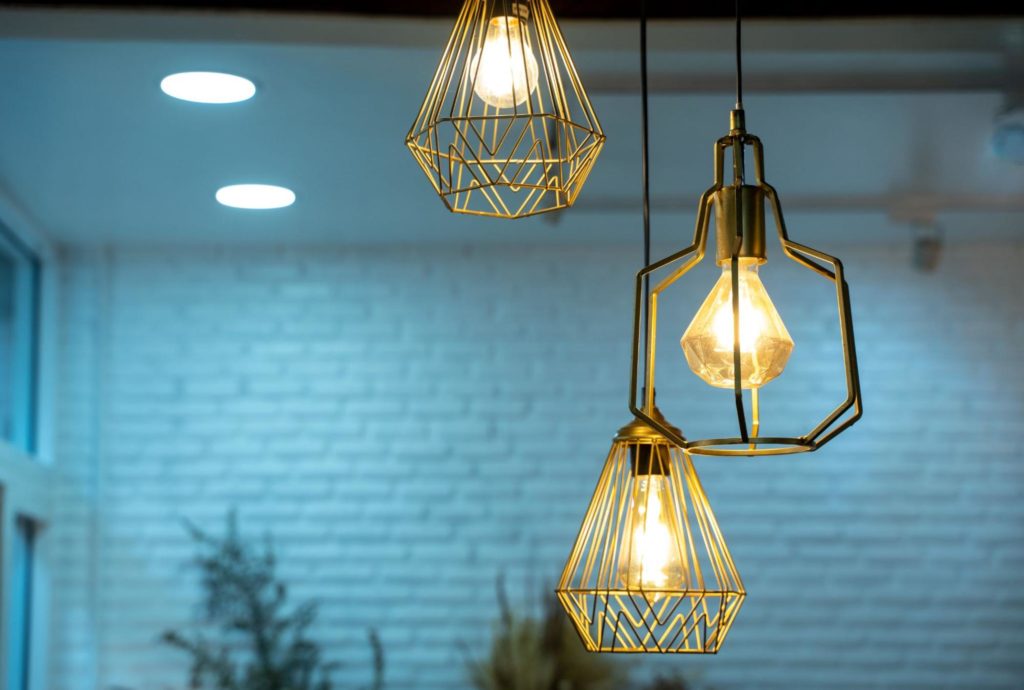 Três luminárias pendentes de metal dourado com lâmpadas expostas, iluminando um ambiente moderno com paredes de tijolos brancos. A luz suave cria uma atmosfera acolhedora e contemporânea.