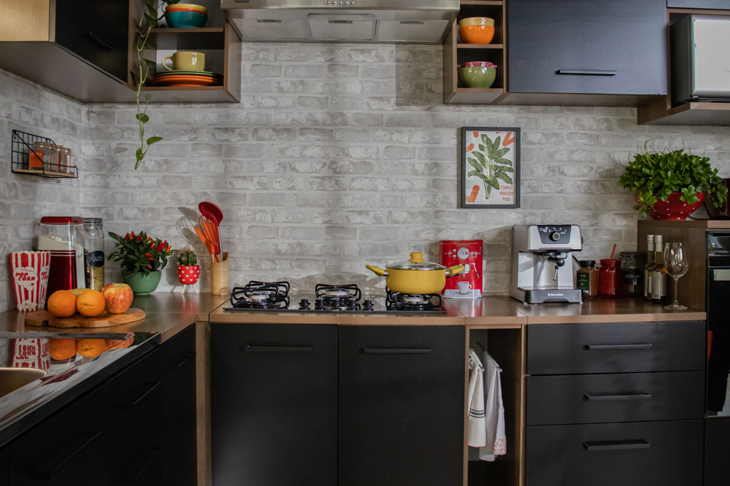 cozinha preta com detalhes em madeira e parede de tijolos na cor cinza conta com objetos decorativos e utensílios coloridos, quadros na parede e vasos de plantas verdes