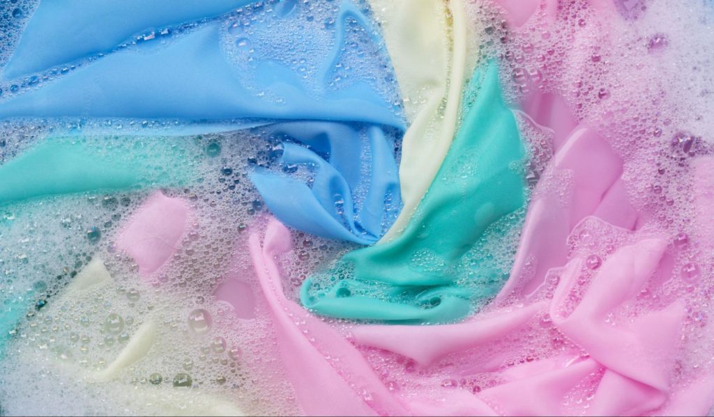 : Close-up de tecidos coloridos em uma lavagem, com bolhas de sabão ao redor.Legenda: Entender como lavar roupa na máquina demanda conhecimento sobre quais cores e tonalidades podem ser misturadas!