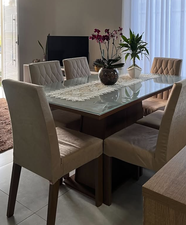 Mesa de jantar de vidro com seis cadeiras estofadas em tecido bege, decorada com um vaso de flores na sala de jantar, ao lado de cortinas brancas.