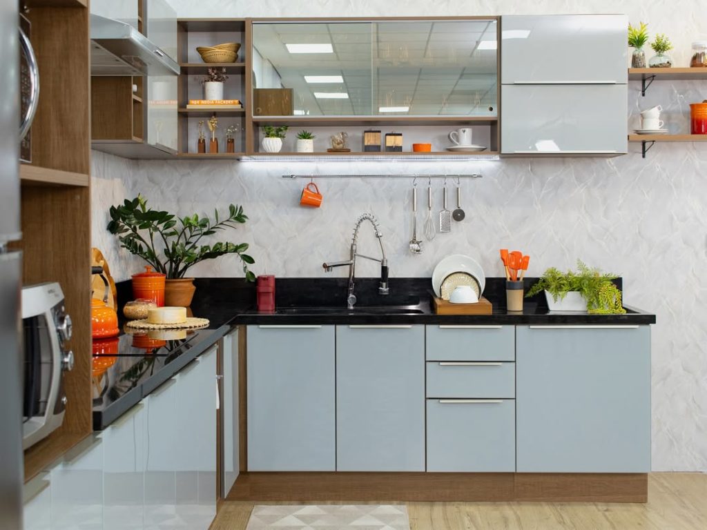 Cozinha moderna com armários cinzas, bancada de granito preto, pia de inox com torneira de cano alto, parede com revestimento branco texturizado, prateleiras de madeira com objetos decorativos, utensílios, plantas e louças na bancada.