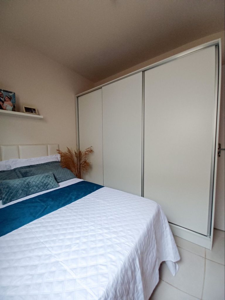 Guarda-roupa branco 100% MDF, com 3 portas de correr, ao lado de uma cama de casal com colcha branca e detalhes em tons de azul.