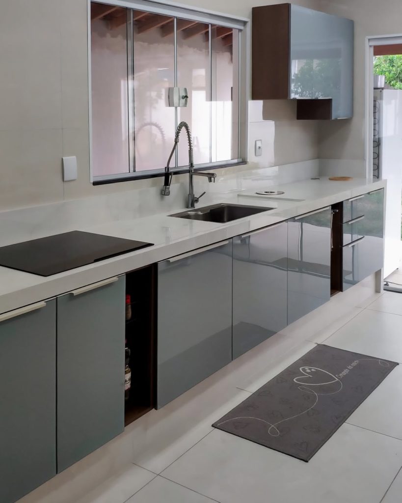 Cozinha com balcão de granito branco, armários de cor cinza com acabamento brilhante, portas espelhadas e uma janela ao fundo.